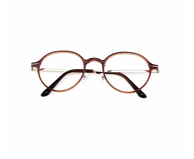 May include: Round eyeglasses featuring a translucent brown plastic frame with thin silver-toned metal temples. The design includes a bridge, adjustable nose pads, and curved temple tips, providing a clear view of the circular lens openings.