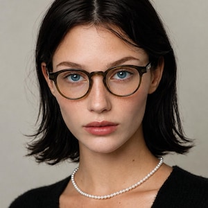 May include: Round olive green acetate eyeglasses with a keyhole bridge and clear lenses. The frames feature a smooth, polished finish and are worn with a simple white pearl necklace and a black textured top, creating a classic and neutral look.