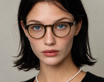 May include: Round olive green acetate eyeglasses with a keyhole bridge and clear lenses. The frames feature a smooth, polished finish and are worn with a simple white pearl necklace and a black textured top, creating a classic and neutral look.