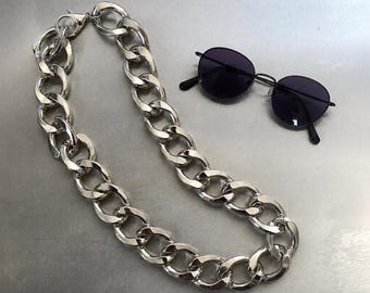 May include: A thick, polished silver-tone metal chain necklace with large, interlocking oval links, positioned alongside a pair of black-framed sunglasses featuring dark, round lenses and thin metal temples.