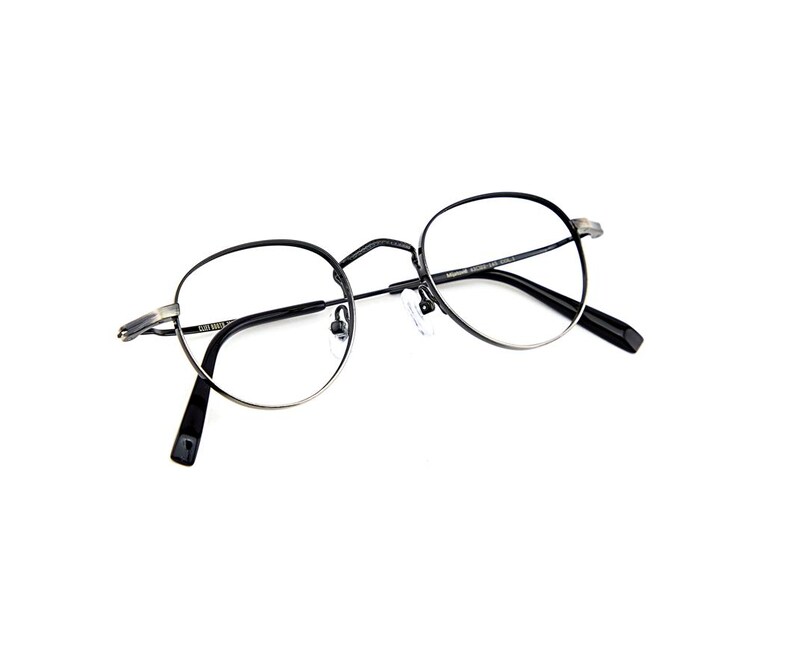 May include: Round eyeglasses featuring a thin, dark metallic frame with a silver-toned finish on the outer edges. The glasses have clear lenses, a bridge, adjustable nose pads, and black temple tips. Text is printed on the inner temple arms.