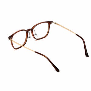 K&ouml;nnte beinhalten: Braune Brille mit rechteckigem Rahmen und goldfarbenen B&uuml;geln. Die Gl&auml;ser sind klar. Die Brille ist leicht angewinkelt, um das Design hervorzuheben. Der Rahmen hat eine warme braune Farbe.
