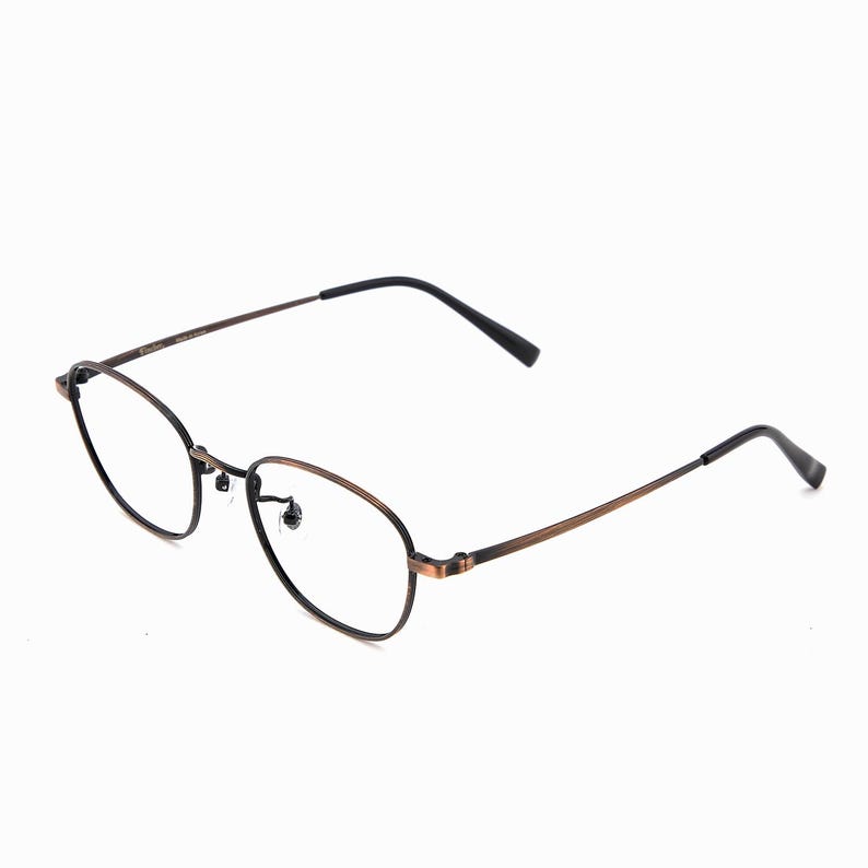 May include: Oval spectacles with a bronze-coloured frame and black temples. The lenses are clear, and the glasses have a classic, minimalist design. The frame is made of metal.
