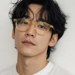 Puede incluir: Una persona con gafas de montura transparente y un tinte amarillo claro. Las gafas tienen forma cuadrada. La persona tiene el pelo oscuro y lleva una camiseta blanca, sobre un fondo blanco.