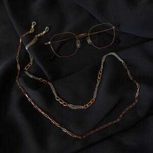 May include: A pair of gold-toned metal eyeglasses with thin frames and a decorative eyeglass chain. The chain features a mix of gold-colored elongated oval links and sections of fine silver-toned metallic mesh, ending in clear rubber loops.