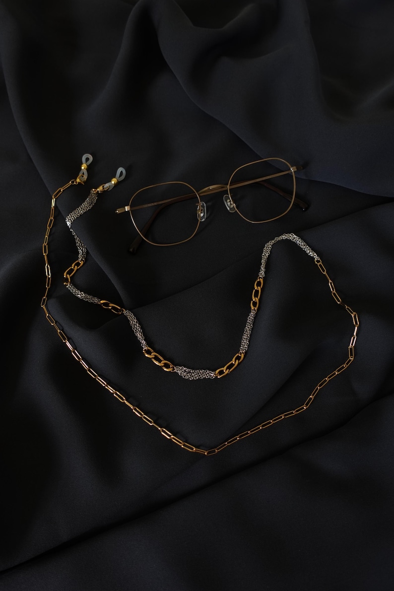 May include: A pair of gold-toned metal eyeglasses with thin frames and a decorative eyeglass chain. The chain features a mix of gold-colored elongated oval links and sections of fine silver-toned metallic mesh, ending in clear rubber loops.