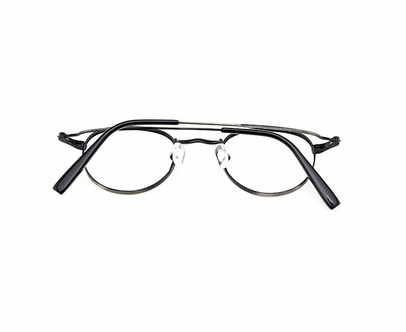 May include: A pair of eyeglasses featuring a thin, dark metallic frame with a double bridge design. The glasses have clear lenses, adjustable transparent nose pads, and black temple tips. The frame has a rounded shape with a sleek, minimalist construction that highlights the structural details of the bridge and the hinges connecting the temples to the front of the eyewear.