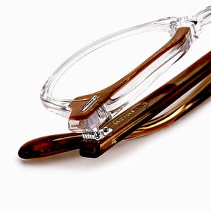 May include: Close-up of eyeglasses with a clear frame and brown tortoiseshell arms. The glasses have a modern design with a visible hinge and the brand name printed on the arm. The lenses are not visible.