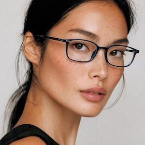 May include: A portrait of a person wearing rectangular, navy blue eyeglasses. The glasses have a thin frame and are positioned on the bridge of the nose. The person has dark hair pulled back, freckles, and a black top.
