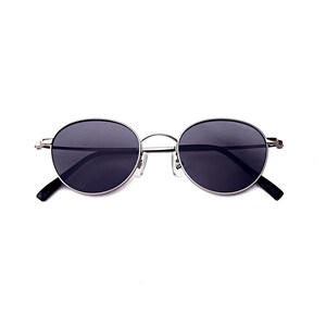 May include: Oval sunglasses with dark lenses and a silver metal frame. The sunglasses have a thin frame and black arms. The lenses are a dark grey colour, and the overall design is minimalist and modern.