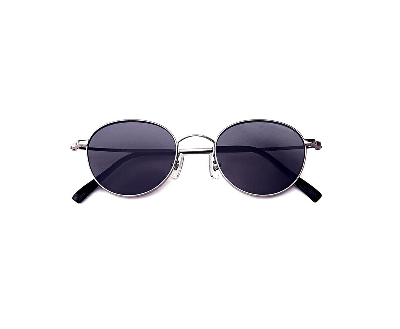 May include: Oval sunglasses with dark lenses and a silver metal frame. The sunglasses have a thin frame and black arms. The lenses are a dark grey colour, and the overall design is minimalist and modern.