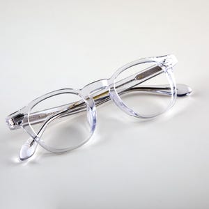 K&ouml;nnte beinhalten: Klare Brille mit rundem Rahmen und transparentem Gestell und B&uuml;geln. Die Brille hat ein klassisches Design mit einem Schl&uuml;ssellochsteg und einem dezenten, rechteckigen Scharnier. Die Gl&auml;ser sind klar, und die Gesamt&auml;sthetik ist modern und minimalistisch.