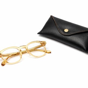 Puede incluir: Un par de gafas con montura transparente y un estuche de cuero negro a juego. Las gafas tienen forma rectangular y est&aacute;n colocadas junto al estuche cerrado. El estuche tiene un cierre de bot&oacute;n dorado.