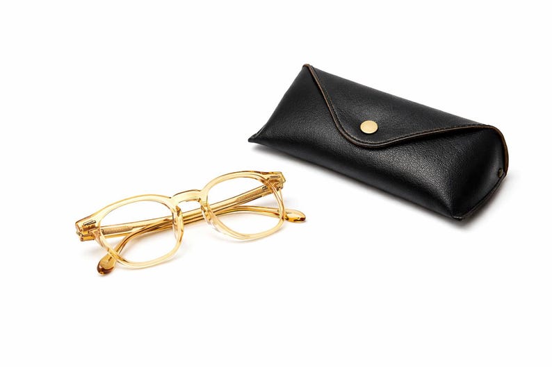 Puede incluir: Un par de gafas con montura transparente y un estuche de cuero negro a juego. Las gafas tienen forma rectangular y est&aacute;n colocadas junto al estuche cerrado. El estuche tiene un cierre de bot&oacute;n dorado.