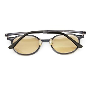 May include: Foldable eyeglasses featuring a translucent dark gray frame and round lenses with a light amber tint. The temples are black, and the nose pads are clear. The glasses are shown in a folded position, highlighting the hinge mechanism.