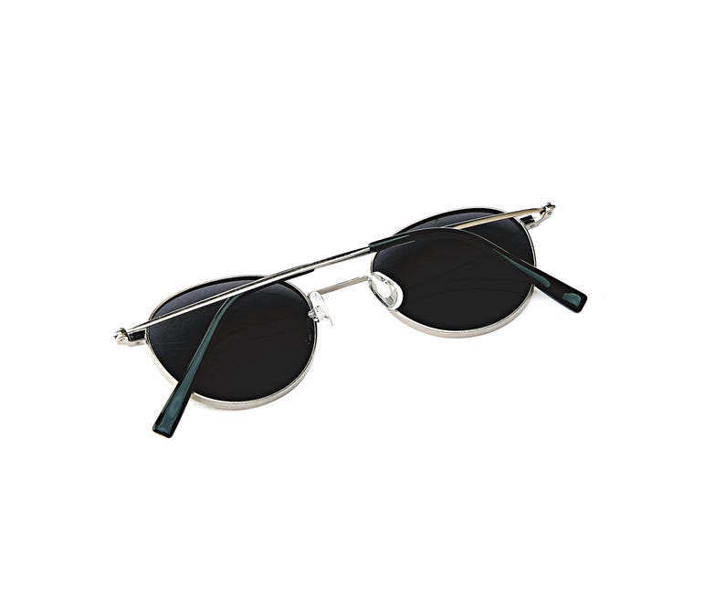 May include: Oval sunglasses with dark lenses and a silver metal frame. The temples are thin and silver, with dark green tips. The sunglasses are set against a white background.