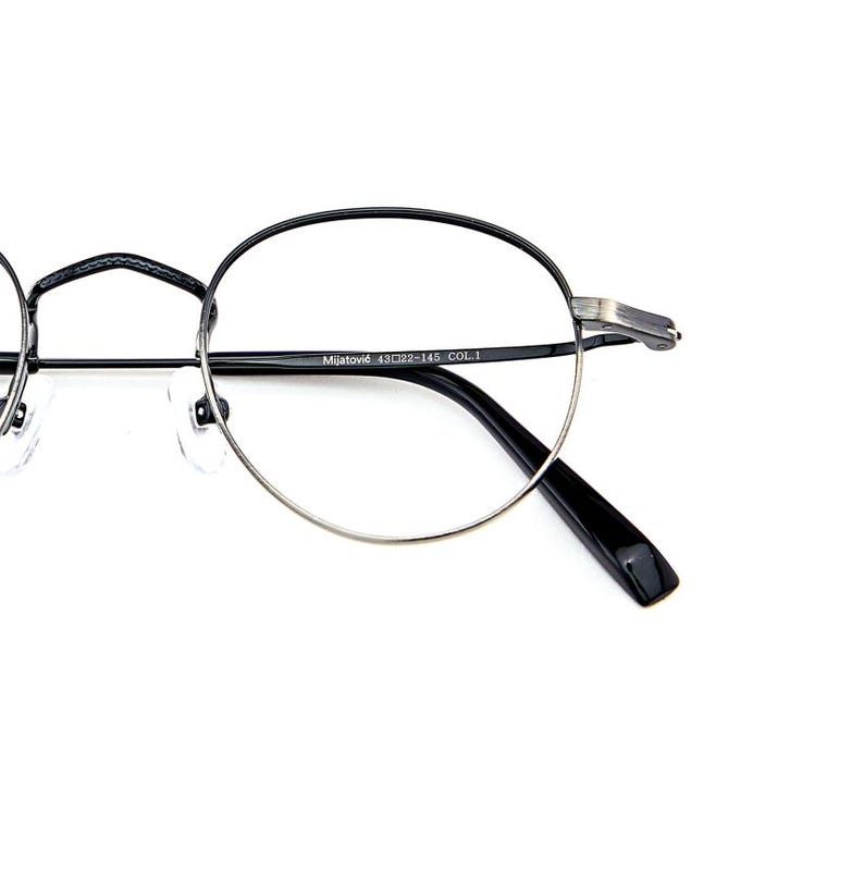 May include: Round eyeglasses with a thin black metal frame and silver-toned lower rims. The inner temple is printed with the text Mijatović 43□22-145 COL.1. The glasses feature clear nose pads and black temple tips, showcasing a sleek design.