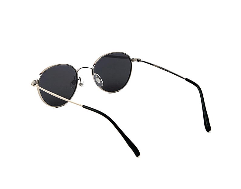 May include: Round sunglasses with dark lenses and a silver frame. The temples are thin and black, with a silver hinge. The sunglasses are set against a white background.