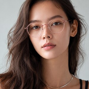 May include: Round eyeglasses with a translucent, pale pink frame and thin metal temples. The glasses feature a classic bridge design with adjustable nose pads, providing a clear view of the circular rims that frame the eyes in a soft, light hue.
