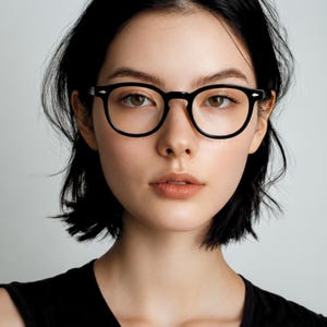 May include: A person with short black hair wearing black-rimmed eyeglasses. The glasses have a classic, rounded shape. The person is wearing a black sleeveless top. The background is a plain, light gray.