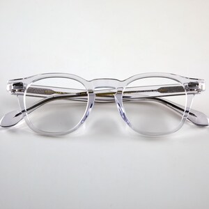 K&ouml;nnte beinhalten: Transparente Brille mit runder Fassung, Schl&uuml;ssellochsteg und dunklen B&uuml;geln. Die Brille besteht aus transparentem Material und bietet eine klare Sicht auf die Gl&auml;ser und die Rahmendetails. Modernes und minimalistisches Design.