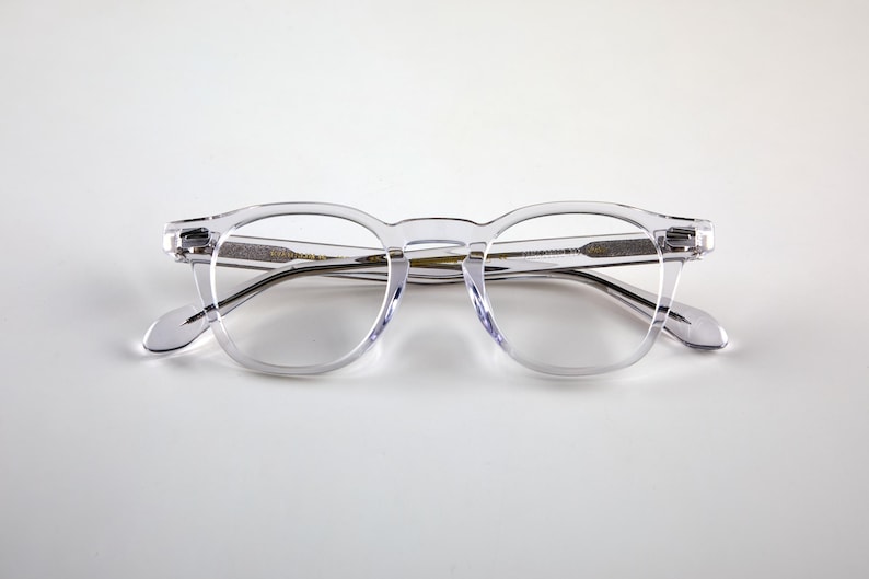 K&ouml;nnte beinhalten: Transparente Brille mit runder Fassung, Schl&uuml;ssellochsteg und dunklen B&uuml;geln. Die Brille besteht aus transparentem Material und bietet eine klare Sicht auf die Gl&auml;ser und die Rahmendetails. Modernes und minimalistisches Design.
