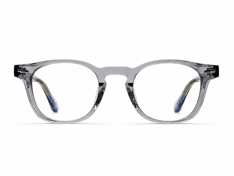 May include: Gray, round-framed eyeglasses with clear lenses. The frames have a translucent appearance, with a subtle gradient on the temples, transitioning from gray to blue. The glasses are set against a plain white background.