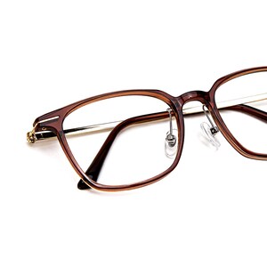 K&ouml;nnte beinhalten: Nahaufnahme einer Brille mit braunem Rahmen und goldfarbenen Akzenten. Die Gl&auml;ser sind klar und die Nasenpads transparent. Die Brille hat eine klassische, rechteckige Form.