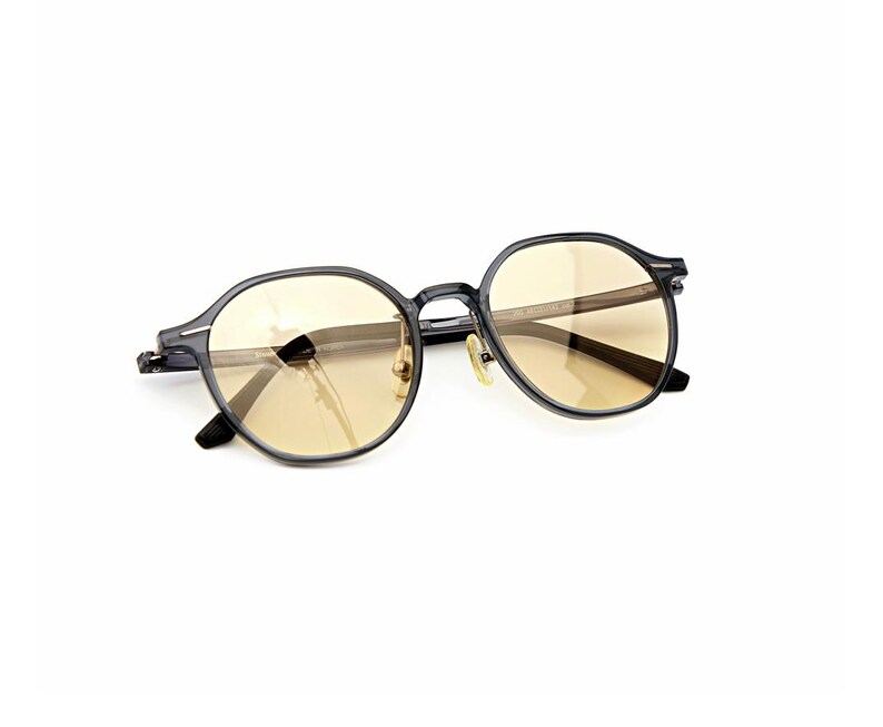 May include: Round eyeglasses featuring a dark translucent grey frame and light amber tinted lenses. The glasses have a thin bridge, metallic nose pads, and slender temples with visible hinges, presenting a clear view of the front and side profile.
