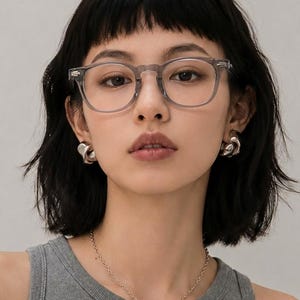 May include: A woman wearing clear-framed eyeglasses, silver earrings, and a silver necklace with a pendant. She is wearing a gray tank top and has short, dark hair.