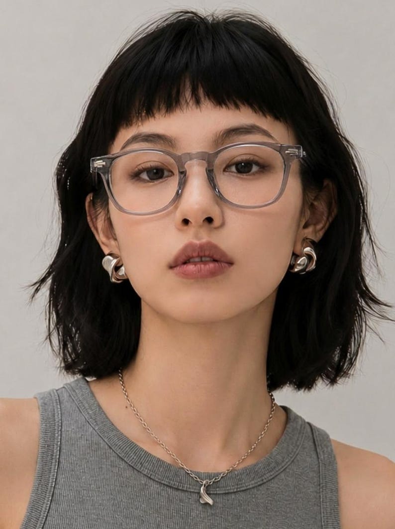 May include: A woman wearing clear-framed eyeglasses, silver earrings, and a silver necklace with a pendant. She is wearing a gray tank top and has short, dark hair.