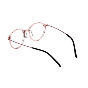 May include: Round eyeglasses featuring a translucent pink frame with thin metallic temples. The temples transition into black plastic tips at the ends. The bridge includes adjustable nose pads, and the overall design maintains a lightweight structure with clear lenses.