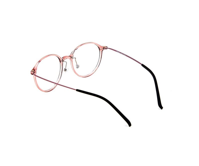 May include: Round eyeglasses featuring a translucent pink frame with thin metallic temples. The temples transition into black plastic tips at the ends. The bridge includes adjustable nose pads, and the overall design maintains a lightweight structure with clear lenses.