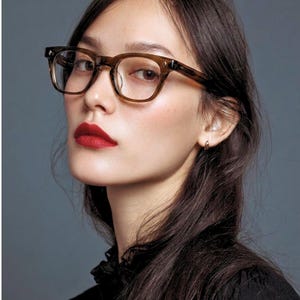 May include: A woman wearing square, brown-framed eyeglasses with clear lenses. She has dark hair, red lipstick, and is wearing a black top. The background is a solid, muted gray.