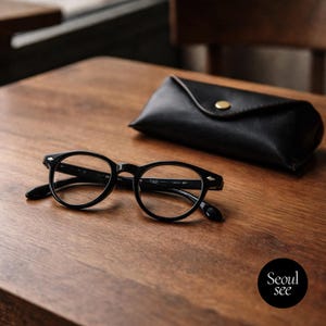 May include: Black-framed eyeglasses with a round lens shape rest on a wooden surface next to a black leather glasses case. The glasses have a classic design. The case is closed with a gold-colored button.
