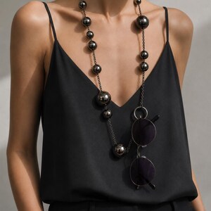 May include: A long necklace featuring polished dark metallic spherical beads of varying sizes strung on a thin chain. A pair of round, dark-tinted sunglasses is suspended from a circular metal ring integrated into the necklace chain.