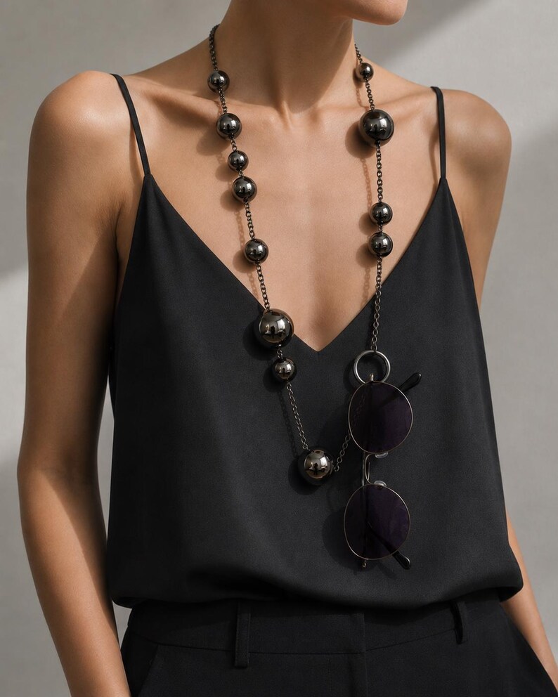 May include: A long necklace featuring polished dark metallic spherical beads of varying sizes strung on a thin chain. A pair of round, dark-tinted sunglasses is suspended from a circular metal ring integrated into the necklace chain.