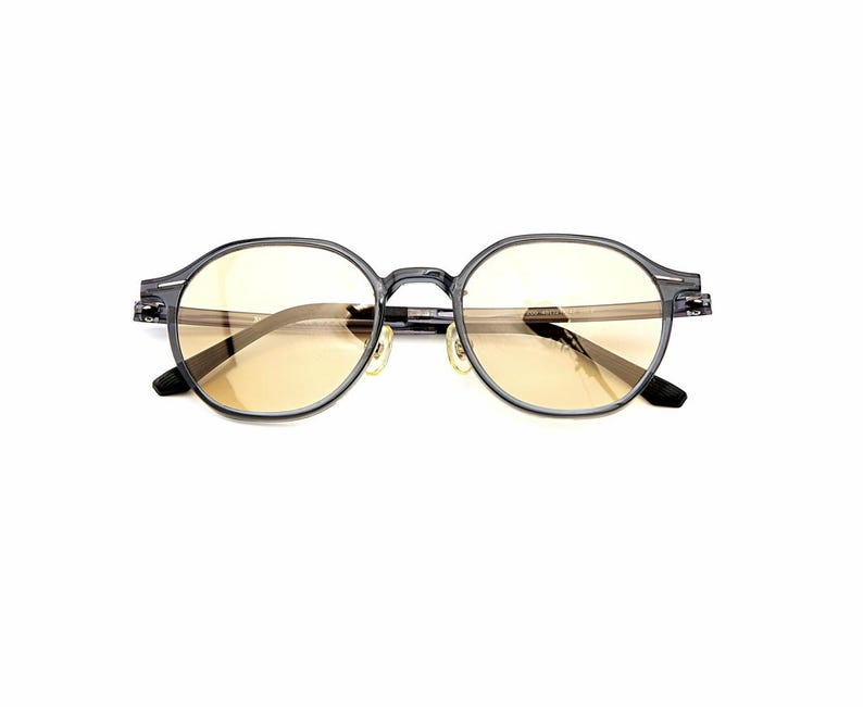 May include: Round-framed glasses featuring a translucent dark gray acetate frame and light amber-tinted lenses. The eyewear includes integrated nose pads and thin temples, showcasing a clear, neutral design suitable for everyday optical wear.