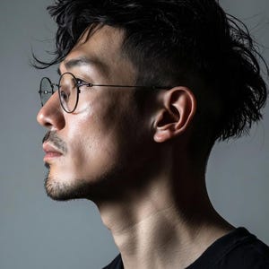 May include: A pair of round-framed glasses with thin black metal rims and clear lenses, worn on a face shown in profile. The glasses feature a minimalist bridge and slender temples that extend behind the ears, providing a clear view of the frame design.