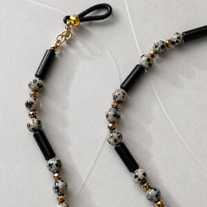 K&ouml;nnte beinhalten: Eine Brillenkette mit einem sich wiederholenden Muster aus schwarzen zylindrischen Perlen, runden Dalmatiner-Jaspis-Perlen mit schwarzen Punkten und kleinen facettierten, metallisch goldenen Zwischenperlen sowie einer schwarzen Gummischlaufe.