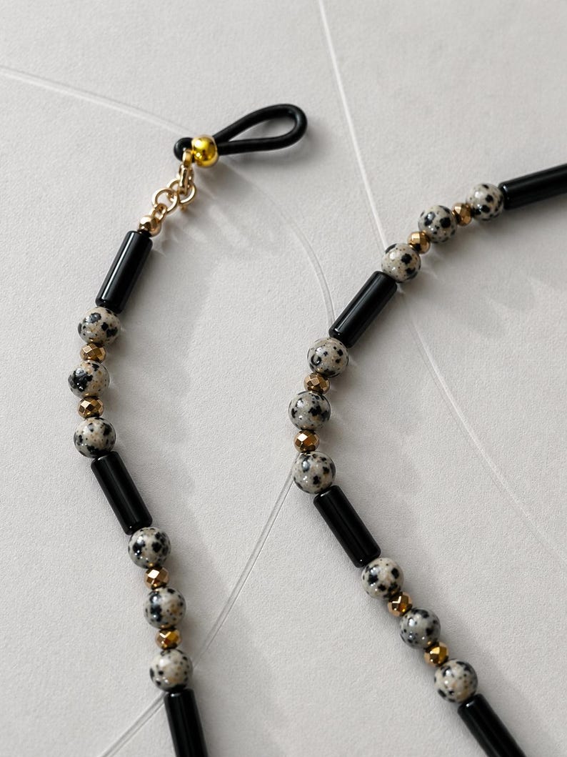 K&ouml;nnte beinhalten: Eine Brillenkette mit einem sich wiederholenden Muster aus schwarzen zylindrischen Perlen, runden Dalmatiner-Jaspis-Perlen mit schwarzen Punkten und kleinen facettierten, metallisch goldenen Zwischenperlen sowie einer schwarzen Gummischlaufe.