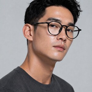 May include: Black round-frame eyeglasses. The glasses have a classic design with a thin frame and are worn by a person. The background is a solid light gray.