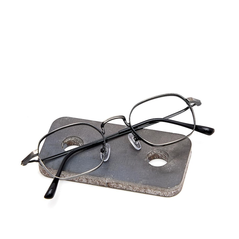 May include: A pair of eyeglasses with thin, dark metallic frames and a rounded, slightly hexagonal shape. The glasses feature clear nose pads and black temple tips, resting on a rectangular, textured gray stone block with two circular holes.
