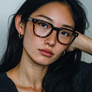 Puede incluir: Una persona con gafas de montura cuadrada con un patr&oacute;n de carey. Las gafas se destacan sobre un fondo de cabello oscuro y piel con pecas. Tambi&eacute;n se ven pendientes de aro dorados.