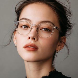 May include: A close-up portrait of a person wearing clear-framed eyeglasses and small gold hoop earrings. The person has brown eyes and light skin. The background is a solid gray.