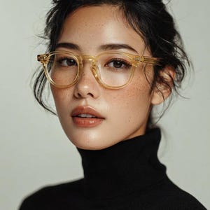 May include: A person wearing clear, square-framed eyeglasses. The glasses have a light yellow tint and are set against a black turtleneck. The person has dark hair and freckles.
