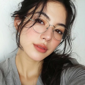 Puede incluir: Un par de gafas de montura met&aacute;lica fina y forma ovalada con acabado plateado. El armaz&oacute;n presenta un puente minimalista y patillas delgadas, sobre un rostro de cabello oscuro que viste una camiseta gris de cuello redondo.