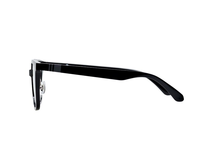 K&ouml;nnte beinhalten: Schwarze rechteckige Brille mit silbernem Scharnier und einem d&uuml;nnen grauen Streifen am B&uuml;gel. Die Brille hat eine gl&auml;nzende Oberfl&auml;che und ist vor wei&szlig;em Hintergrund platziert.