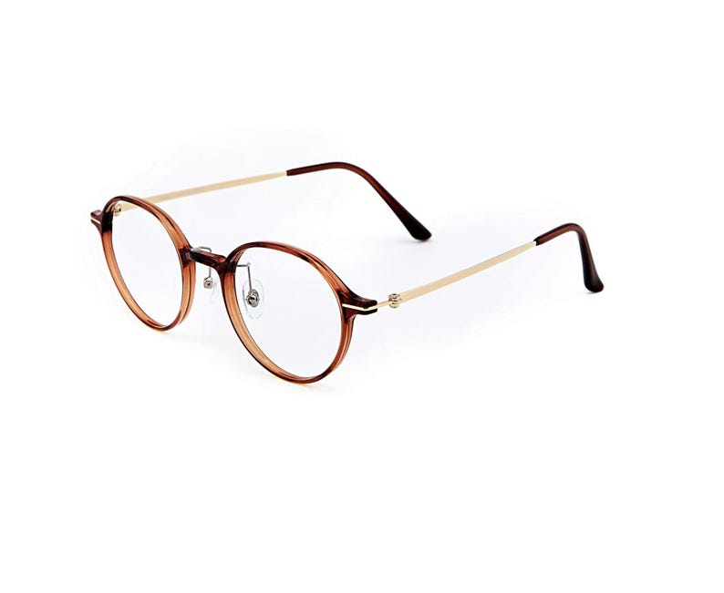 May include: Round eyeglasses featuring a translucent brown acetate frame with a thin gold-tone metal bridge and temples. The temples transition into dark brown acetate tips. The clear lenses are held by a thin rim with adjustable nose pads.