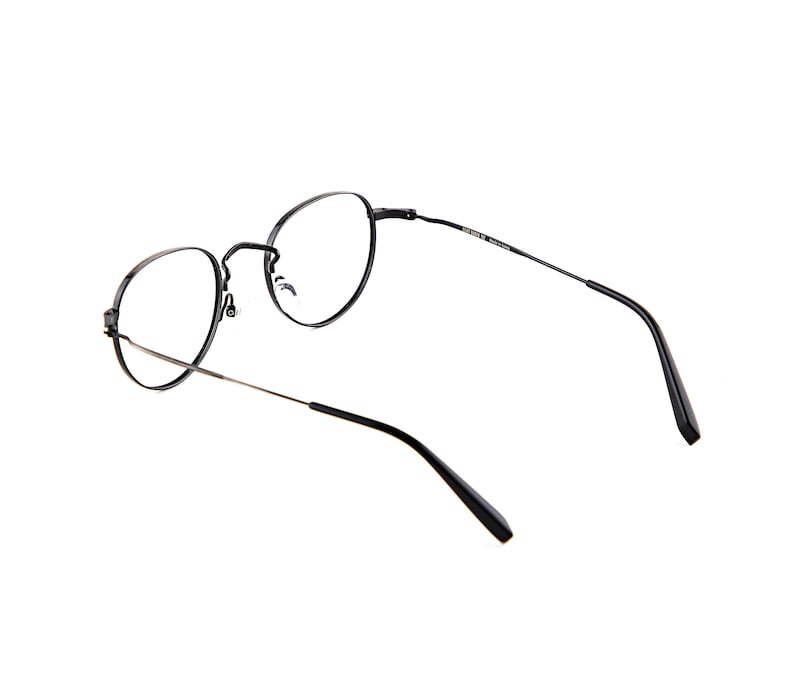 May include: Black metal eyeglasses with thin, circular frames and slender temples. The glasses feature clear lenses, a bridge with nose pads, and black plastic temple tips. The inner side of the temple is marked with text including the brand name.